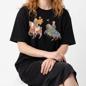 Puede incluir: Camiseta negra con una colorida ilustraci&oacute;n de dos figuras a caballo, adornadas con coronas y s&iacute;mbolos celestiales. El dise&ntilde;o presenta detalles intrincados y una est&eacute;tica vintage. La camiseta es de estilo casual y c&oacute;modo.