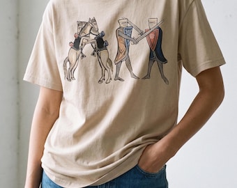 Puede incluir: Una camiseta beige de manga corta con un estampado gráfico de dos caballos encabritados y dos caballeros con armadura de cota de malla en un duelo de espadas. La ilustración tiene un estilo medieval con detalles de escudos azules y rojos.