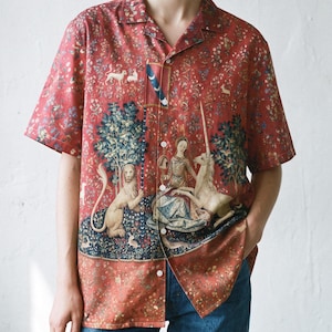May include: A short-sleeved button-down shirt in a deep red hue, featuring a detailed tapestry-style print of a lion, a unicorn, and a figure. The design includes intricate floral patterns, a blue banner with crescent moons, and white buttons.