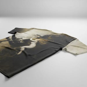 May include: A short-sleeved t-shirt featuring a classical painting of a nude figure with long hair. The artwork is predominantly dark with lighter shades of beige and white, creating a dramatic contrast. The shirt is laid flat on a white surface.