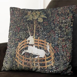 May include: A black and white throw pillow with a tapestry design featuring a white unicorn in a circular fence with a tree behind it. The background is a dark green with a variety of flowers and leaves.