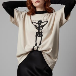 Puede incluir: Camiseta beige con un gr&aacute;fico de esqueleto negro y el texto "VENIR DU CABARET DU NEANT". La camiseta tiene cuello negro y mangas largas. La camiseta est&aacute; hecha de un material suave y c&oacute;modo.
