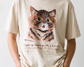 May include: A beige short-sleeve t-shirt featuring a brown tabby kitten illustration with large eyes and an open mouth. Below the kitten, the text reads: Louis Wain. I fell in love with a lovely kitten. That kitten was myself. The shirt has a crew neck.