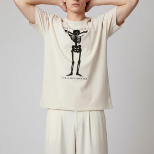 Puede incluir: Camiseta color crema con un dise&ntilde;o de esqueleto negro y el texto "VENIR PAR-DEL&Agrave; DU N&Eacute;ANT" arriba y "PARIS-MONTMARTRE" abajo. La camiseta se combina con pantalones color crema.