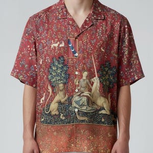 Op de afbeelding: Rood shirt met korte mouwen met een print in tapijtstijl met een leeuw, een eenhoorn en een zittende figuur. Het shirt heeft een kampkraag en een knoopsluiting. De print bevat bloemen- en diermotieven.