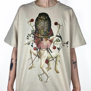 May include: Beige t-shirt featuring a surrealist illustration. The artwork depicts a large owl atop a figure with outstretched arms holding red cherries. The figure is surrounded by plant-like elements and more cherries. The design has a vintage, artistic aesthetic.