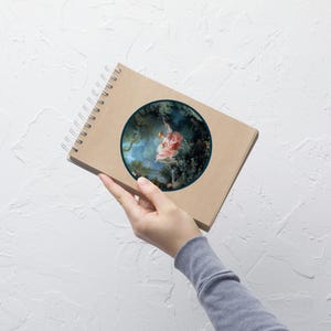 May include: A spiral-bound notebook with a brown cover. The notebook features a circular image of a woman on a swing in a forest. The artwork is framed by a dark green border. The notebook is held by a person.