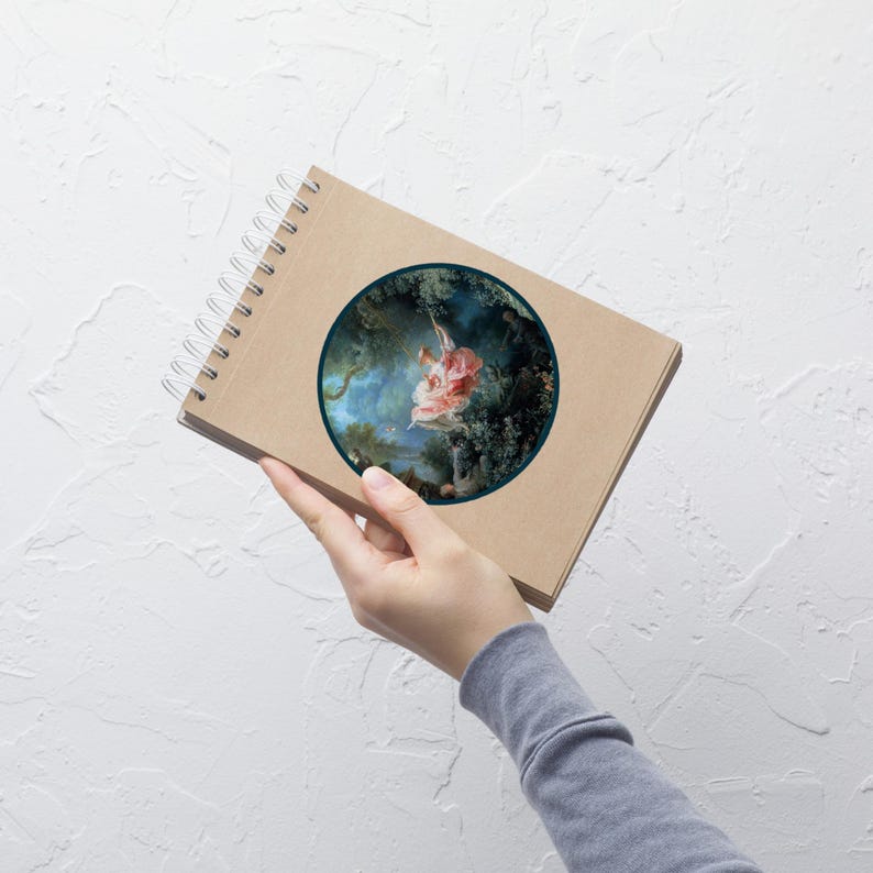 May include: A spiral-bound notebook with a brown cover. The notebook features a circular image of a woman on a swing in a forest. The artwork is framed by a dark green border. The notebook is held by a person.