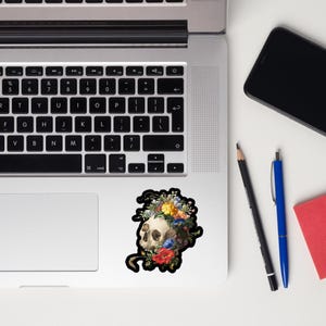 May include: A sticker featuring a skull with a floral crown. The skull is white and the flowers are colorful, including red, yellow, and blue. The sticker is on a laptop.