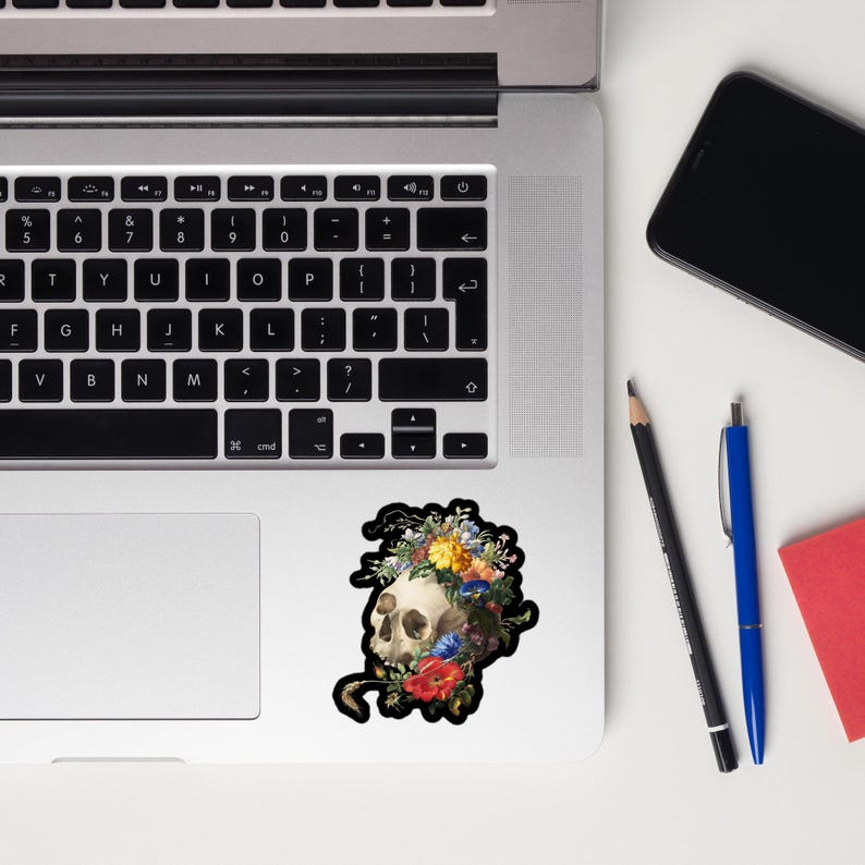 May include: A sticker featuring a skull with a floral crown. The skull is white and the flowers are colorful, including red, yellow, and blue. The sticker is on a laptop.