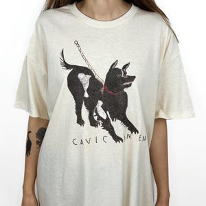 以下が含まれることがあります： クリーム色のTシャツに、鎖につながれた犬の白黒のイラストが描かれています。犬の下には「Cave Canem」という文字が印刷されています。