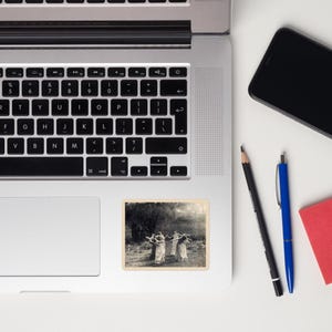 May include: A sticker featuring a black and white image of a group of people dancing in a circle outdoors. The sticker is rectangular with a cream-colored border and is placed on a laptop.