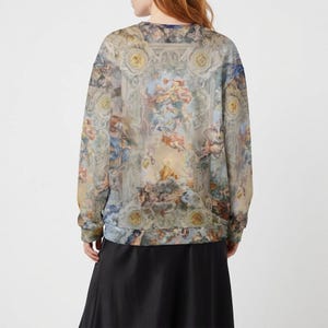 May include: A long-sleeved sweatshirt featuring a Renaissance-style fresco print in shades of blue, beige, and peach. The artwork depicts cherubs, architectural details, and celestial elements. The sweatshirt is paired with a black skirt.