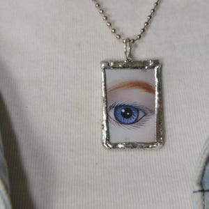 May include: Silver-toned necklace with a rectangular pendant featuring a detailed illustration of a blue eye. The eye has a light skin tone and reddish-brown eyebrows. The pendant is framed with a silver border and hangs from a ball chain.