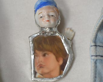 Può includere: Una collana con catena d'argento con un ciondolo di porcellana a forma di bambola. La bambola indossa un cappello blu e ha una foto di un viso umano all'interno del suo petto.