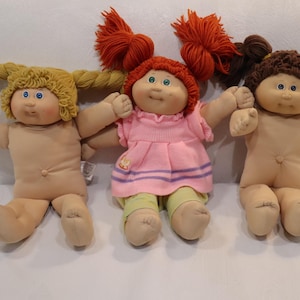May include: Three Cabbage Patch Kids dolls with yarn hair. The left doll has blonde braided hair, the center doll has red pigtails and wears a pink dress with yellow pants, and the right doll has brown pigtails. All dolls have blue eyes.