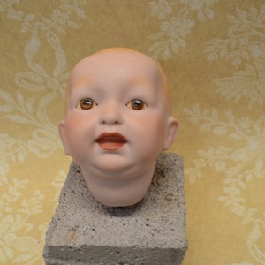 May include: A porcelain doll head with brown eyes and a light pink complexion. The head is mounted on a gray concrete block.