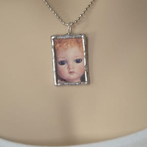 May include: A rectangular pendant featuring a printed image of a doll face with blue eyes and curly blonde hair, encased in a silver-toned metal frame with a textured edge, suspended from a silver-toned ball chain necklace.