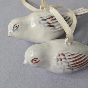 May include: Two white ceramic bird ornaments with brown wing details and red eyes. Each bird has a cream-colored ribbon for hanging. The ornaments are likely for holiday or home decor.