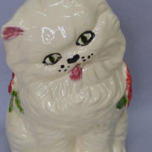 May include: A ceramic figurine of a white cat with green eyes, black nose and whiskers, and a pink tongue sticking out. The cat has red accents on its ears and a red bow with green leaves around its neck. The figurine is sitting upright.