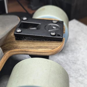 Könnte beinhalten: Nahaufnahme eines Skateboard-Decks aus Holz mit schwarzer Metallbeschläge und einem Logo. Das Deck ist auf zwei weißen Rädern montiert.