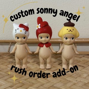 Może przedstawiać: Trzy niestandardowe figurki Sonny Angel. Figurki są nagie z różnymi nakryciami głowy: białym kapeluszem Hello Kitty z czerwoną kokardą, czerwoną czapką z białym kwiatem i żółtym kapeluszem z brązową górą. Tekst brzmi "custom sonny angel" i "rush order add-on".