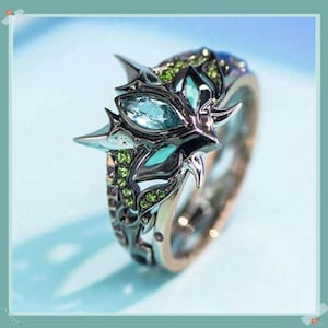Könnte beinhalten: Ein detaillierter Silberring mit einzigartigem Design. Der Ring hat einen zentralen hellblauen Edelstein, umgeben von kleineren grünen Steinen und aufwendigen Metallarbeiten. Das Band hat eine goldfarbene Innenseite.