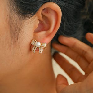 May include: Gold-toned bow-shaped stud earrings with a central pearl and sparkling clear crystals. The earrings are a delicate design, suitable for adding a touch of elegance to any outfit. The earrings are shown on an ear.