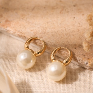 Puede incluir: Un par de pendientes de aro dorados con grandes perlas color crema colgantes. Los pendientes se exhiben sobre una superficie beige texturizada, con un fondo suave y neutro.