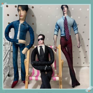 Puede incluir: Tres muñecos de tela hechos a mano. Uno lleva una chaqueta vaquera y vaqueros, otro un traje negro y gafas de sol, y el tercero una camisa azul claro, pantalones rojos y una corbata a rayas. Los muñecos están colocados delante de un panel perforado blanco.