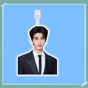 Puede incluir: Una pegatina con un hombre con traje negro, camisa blanca y corbata negra. La pegatina tiene un borde blanco y un colgador de plástico transparente en la parte superior. El fondo es de color azul claro.