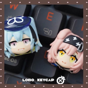 Peut inclure: Deux touches de clavier artisanales personnalisées représentant des têtes de personnages de style anime. La touche de gauche a des cheveux bleu clair et un béret bleu foncé. Celle de droite a des cheveux roses et un bandeau noir avec des éclairs blancs. Texte : LOBO KEYCAP.