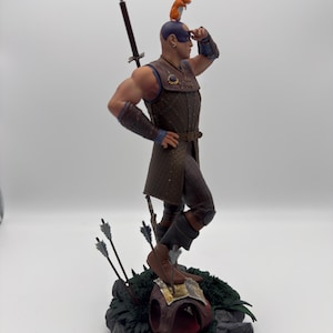 May include: A detailed figurine of a warrior with a sword and arrows, standing on a rocky base with green foliage. A small orange animal sits on the warrior's head.