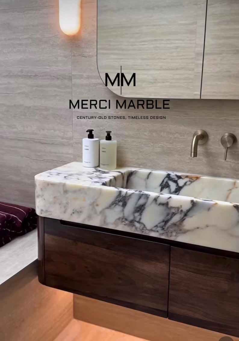 May include: A modern bathroom vanity with a marble countertop and dark wood cabinetry. The countertop features white and gray veining. Two white bottles sit on the counter. The text "MERCI MARBLE" is visible.