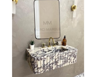 May include: A white and purple marble bathroom sink with a gold faucet and a black framed mirror above it. The mirror has the text "MM Merci Marble" on it.