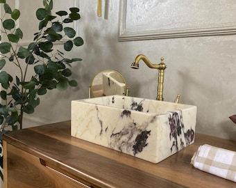 May include: A white marble bathroom sink with purple and brown veins sits on a wooden countertop. A gold faucet is attached to the sink.