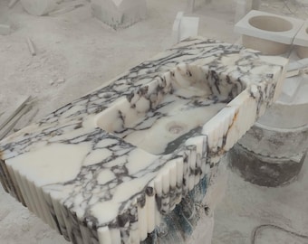 May include: A rectangular marble sink with a built-in basin. The sink features a white base with dark gray veining and a textured, vertically-lined edge. The sink is in a workshop setting, surrounded by other stone works.