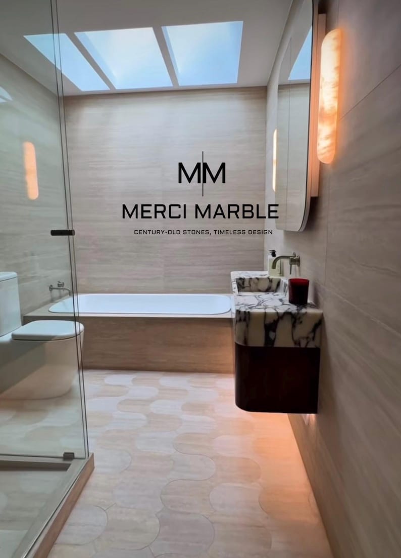 May include: A modern bathroom featuring a bathtub, toilet, and vanity with marble accents. The room has a neutral color palette with a skylight and the text "MERCI MARBLE CENTURY-OLD STONES, TIMELESS DESIGN".