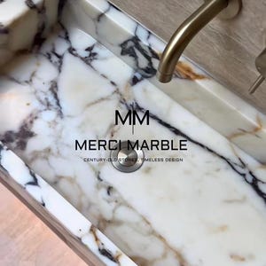 May include: A white marble sink with black and brown veining. The sink has a brushed gold faucet and drain. The text "MM MERCI MARBLE CENTURY-OLD STONES, TIMELESS DESIGN" is printed on the sink.