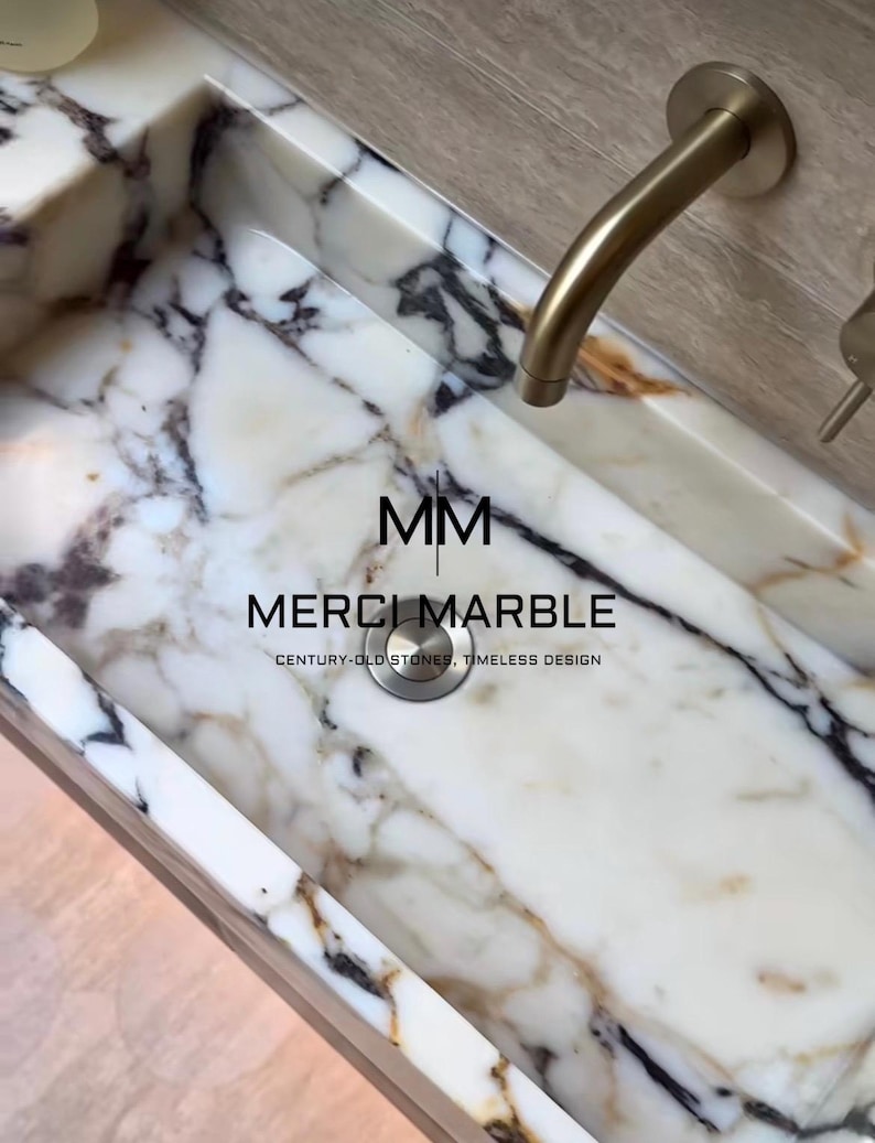 May include: A white marble sink with black and brown veining. The sink has a brushed gold faucet and drain. The text "MM MERCI MARBLE CENTURY-OLD STONES, TIMELESS DESIGN" is printed on the sink.