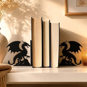 Könnte beinhalten: Ein Paar Buchstützen aus schwarzem Metall mit der Silhouette eines auf einem Felsen sitzenden Drachen. Die Buchstützen stehen auf beiden Seiten von drei Hardcover-Büchern mit cremefarbenen Seiten und halten diese in aufrechter Position.