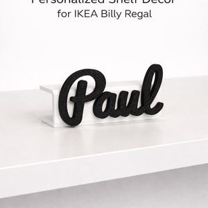 Może przedstawiać: Czarna, pokryta brokatem nazwa „Paul” znajduje się na białej półce. Półka jest elementem dekoracyjnym do regału IKEA Billy. Tekst „Personalized Shelf Decor for IKEA Billy Regal” znajduje się u góry obrazu.