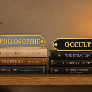 Może przedstawiać: Dwa dekoracyjne znaki, jeden z napisem "PHILOSOPHIE" złotymi literami na szarym tle, a drugi z napisem "OCCULT" w kolorze złotym na czarnym tle. Znaki znajdują się na stosach książek z tytułami takimi jak "The Republic" i "The Kybalion".