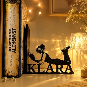 May include: A black metal bookend featuring a silhouette of a person lying down while reading a book, with the name KLARA cut out in capital letters at the base. The design is positioned next to several books on a shelf in a warm, dimly lit setting.