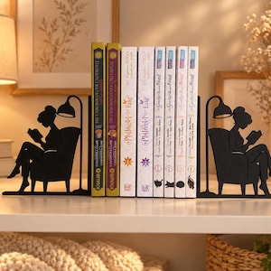 Op de afbeelding: Een paar boekensteunen van zwart metaal met het silhouet van een persoon die in een fauteuil onder een vloerlamp een boek leest. De boekensteunen houden een rij van zeven boeken vast met diverse titels waaronder Philome the Fake.