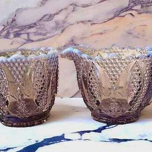 May include: A vintage, iridescent glass creamer and sugar bowl set. The pieces have a diamond pattern and a scalloped rim. The creamer has a handle. The set is a soft purple color with hints of blue and sits on a white surface.