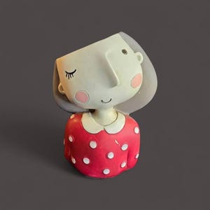 May include: A whimsical ceramic planter shaped like a stylized girl's head. The face is off-white with closed eyes, rosy cheeks, and a small smile. The red dress is adorned with white polka dots and a white collar. A unique decorative item.