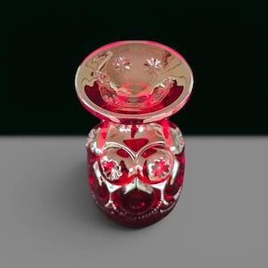 May include: A vibrant ruby red glass vase featuring a wide, flared rim and a rounded body. The surface is decorated with a repeating pattern of raised circular medallions, each containing a star-like embossed detail, creating a textured finish.