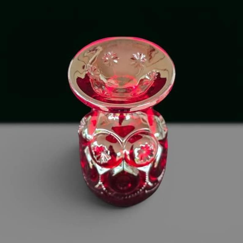 May include: A vibrant ruby red glass vase featuring a wide, flared rim and a rounded body. The surface is decorated with a repeating pattern of raised circular medallions, each containing a star-like embossed detail, creating a textured finish.