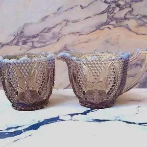 May include: A vintage glass sugar bowl and creamer set. The pieces are a lavender color with a diamond and floral pattern. The creamer has a handle and spout. The set is displayed on a white surface with a marble background.